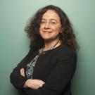 Smiling woman with curly dark hair and glasses, arms crossed, black blazer, teal backdrop