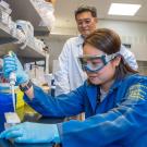 Focused woman pipetting in lab, blue coat, gloves, goggles; male colleague watching