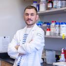 Smiling young man in white lab coat, arms crossed, in chemical lab with shelves of bottles