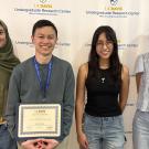 Four smiling students before a university logo backdrop, one holding a certificate