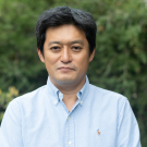 Man in light-blue button-down shirt standing before blurred green foliage, neutral expression