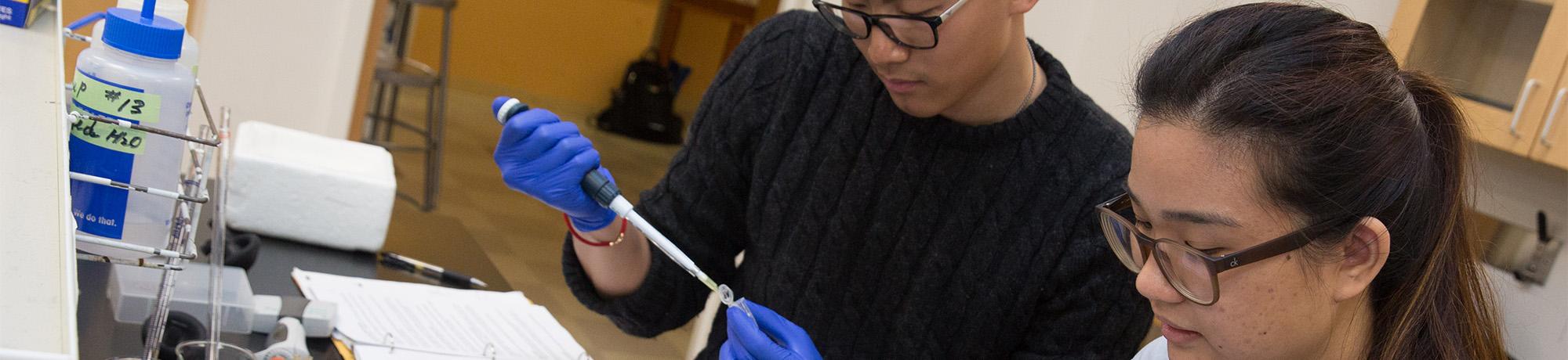 Two gloved researchers concentrating at a lab bench, one using a pipette