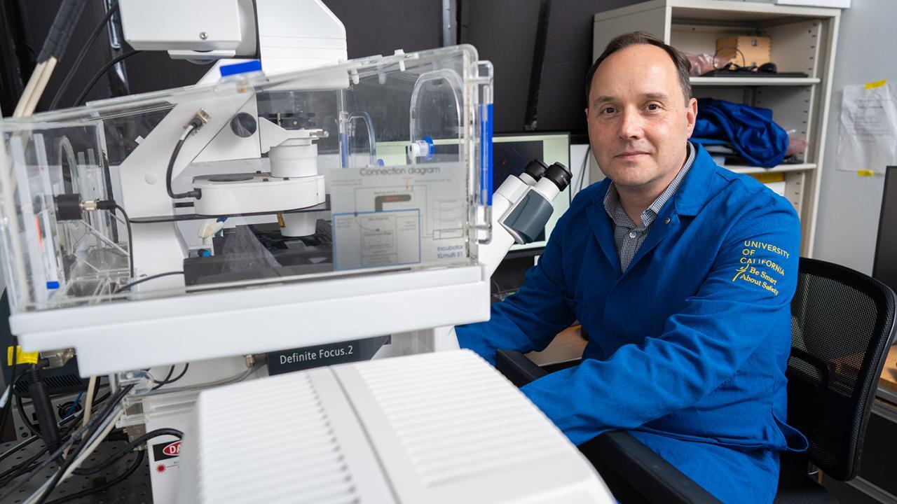 Researcher in a UC lab coat sits beside advanced microscope equipment, preparing for imaging work in the laboratory.
