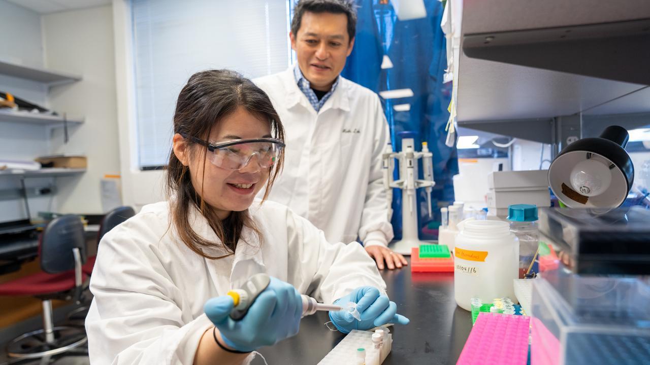 A young female scientist in a lab coat and goggles pipettes a sample while a male scientist observes in a well-equipped laboratory.